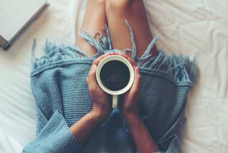 A person is lying on a bed with their legs up, holding a cup of coffee and a book.