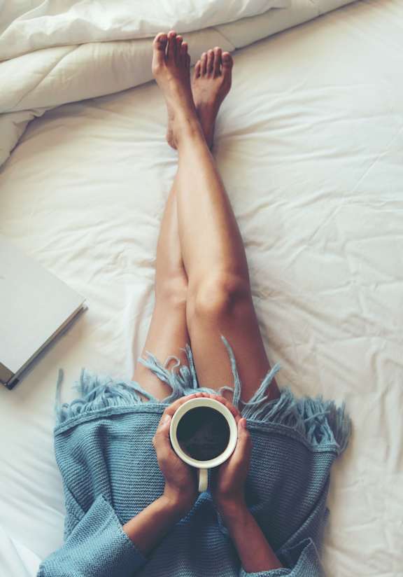 A person is lying on a bed with their legs up, holding a cup of coffee and a book.
