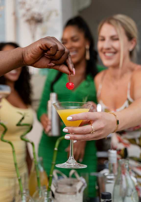 A person is holding a glass with a yellow drink and a cherry on top.