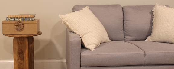 A grey couch with a white pillow and a wooden side table with a box on top.
