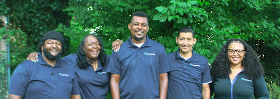 A group of five people wearing blue shirts stand in front of a green tree.