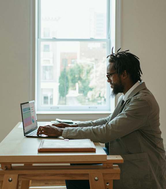 A man with dreadlocks is working on a laptop at a wooden desk.