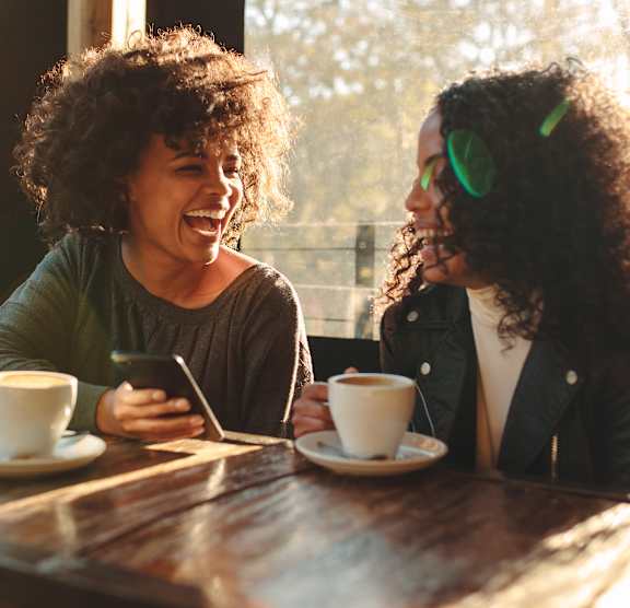 Two people sitting at a table with cups of coffee and one person holding a phone.