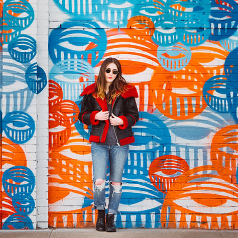 A woman in a red and black jacket and ripped jeans stands in front of a wall with blue and orange artwork.