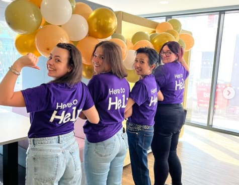 Four women wearing purple shirts with the word "Hero" on them are standing in front of a balloon arch.
