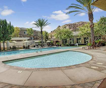 The turquoise outdoor pool with pool chairs, tables, and chairs with the apartments and the desert hills in the background at Rancho Belago Apartments. 