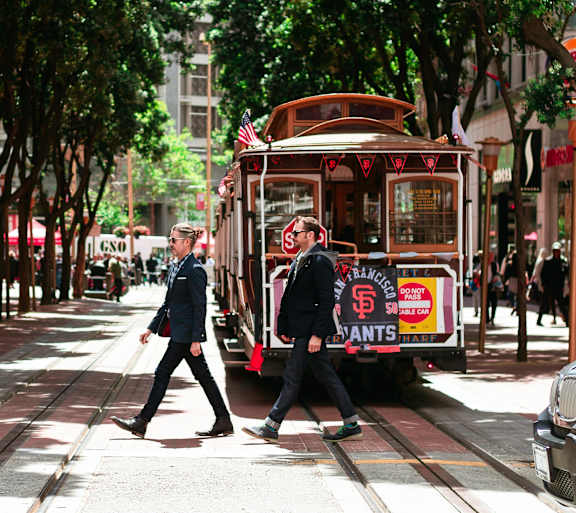 Two men are walking on a street with a cable car in the background.