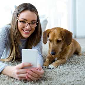 A woman with glasses is lying on the floor with a dog, looking at a tablet.