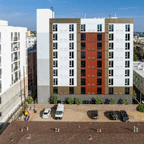 A large multi-story building with a red and white facade is surrounded by other buildings and has cars parked in front.