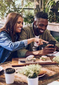 A man and a woman are sitting at a table with a smartphone in the man's hand.