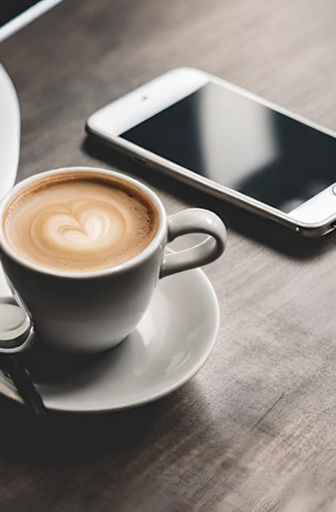 A cup of coffee with a heart design on top sits on a saucer next to a smartphone.