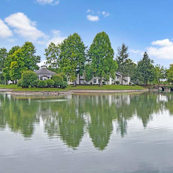 The exterior of the apartments and the green space with grass, trees, shrubs, and a pond with a bridge at Island Park. 