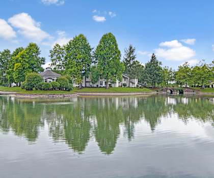 The exterior of the apartments and the green space with grass, trees, shrubs, and a pond with a bridge at Island Park. 