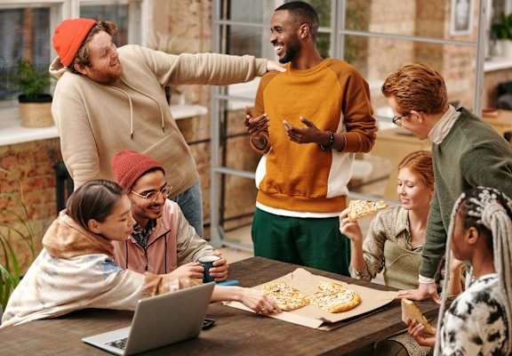 A group of people are gathered around a table with a laptop and pizza, seemingly engaged in a casual meeting or discussion at Crogman School Lofts in GA, 30315