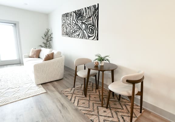 A living room with a white couch, a small table with two chairs, and a large abstract painting on the wall.