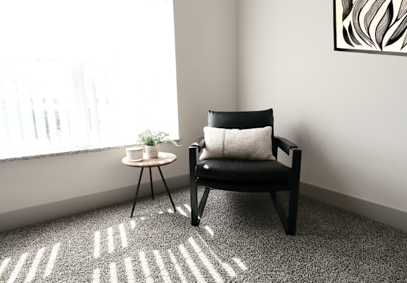 A black chair with a white pillow sits in front of a window with a small table beside it.