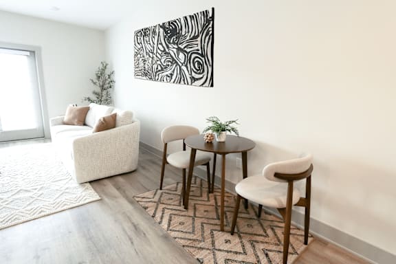 A living room with a white couch, a small table with two chairs, and a large abstract painting on the wall.