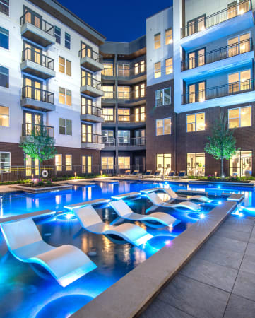 Courtyard pool and tanning area during night time with bright lights in water and view of surrounding apartments and balconies