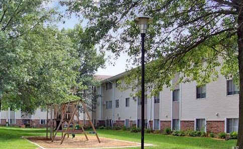 A playground with a swing set is in the foreground of a grassy area in front of apartment buildings.at Foxwood Apartments, Indianola, IA
