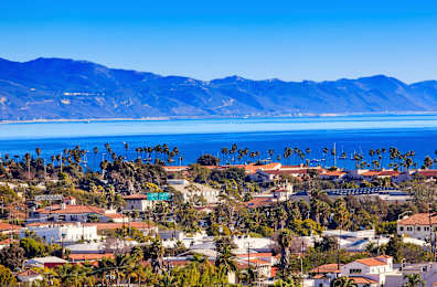 A coastal town with houses and palm trees in the foreground and mountains in the background.