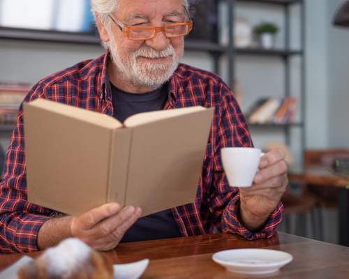 An elderly man is reading a book while holding a cup.