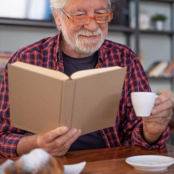 An elderly man is reading a book while holding a cup.