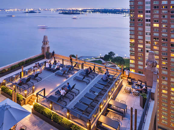 A rooftop bar overlooks a body of water with boats and a city skyline in the distance.  at One West, New York, New York