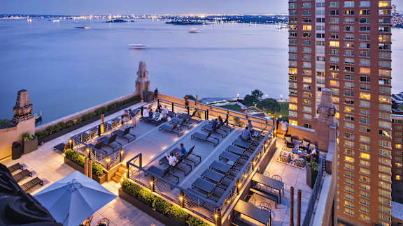 A rooftop bar overlooks a body of water with boats and a city skyline in the distance.  at One West, New York, New York