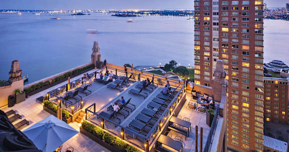 A rooftop bar with a view of the water and boats at dusk.