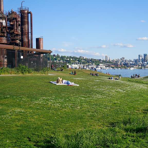 A grassy field with people sitting and relaxing with a city skyline in the background.