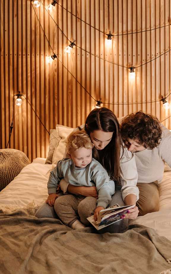 A family of three is sitting on a bed with a book, with string lights hanging above them.