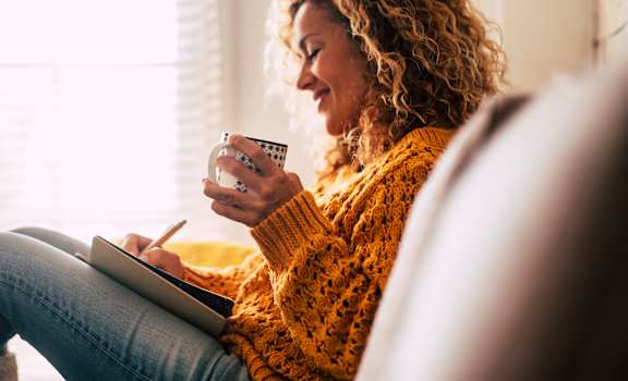 A woman with curly hair is sitting on a couch, writing in a notebook and holding a mug.