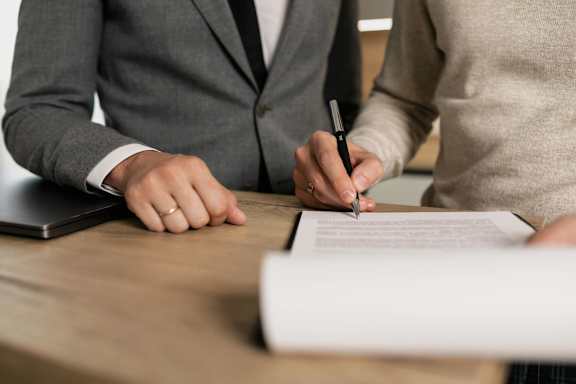 A person in a suit is signing a document while another person holds a pen.
