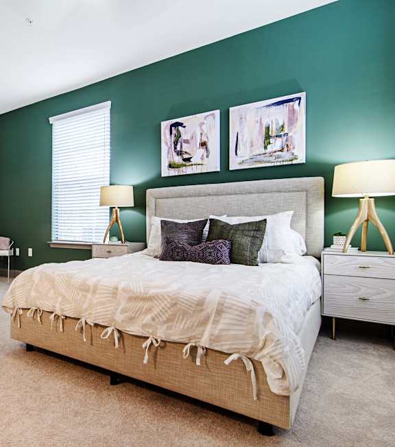 An interior view of a bedroom with a bed, two side tables with lamps on them, and a window at Parkstone at Knightdale apartments in Knightdale, NC.