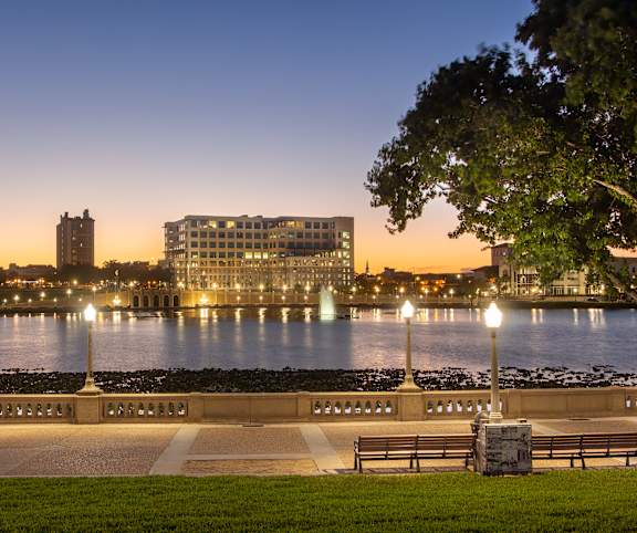 A park with a bench and lamp posts overlooking a body of water with buildings in the background.
