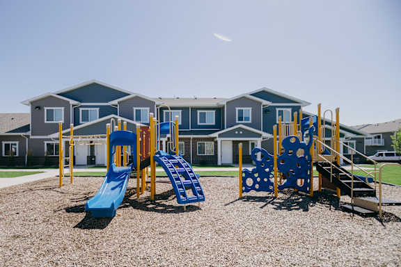 A playground with a blue slide and yellow and blue swings in front of a building.