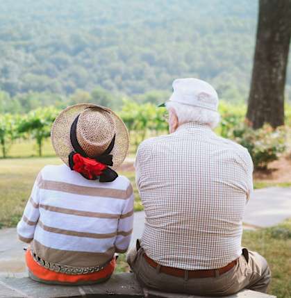 An elderly couple sits together on a stone bench looking out over a lush green landscape.