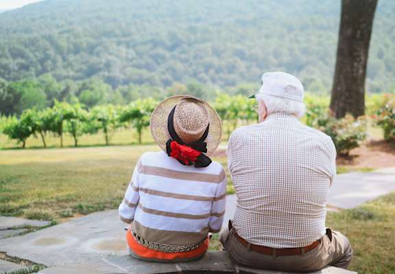 An elderly couple sits together on a stone bench, looking out over a lush green landscape.