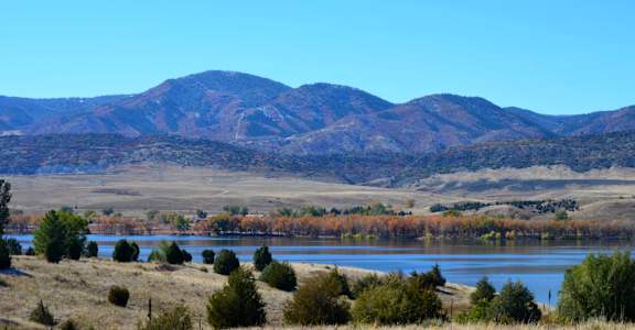 A mountain range with a lake in the foreground and autumn trees in the middle ground.