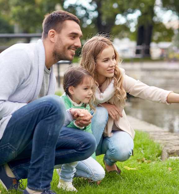 A family of three is sitting on the grass by a pond.