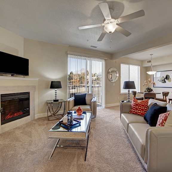 The spacious, bright living room with a couch, throw pillows, large windows, a ceiling fan, wall art, plush chairs, coffee table, decorations, a TV, and glass doors to the balcony at Vista View Apartment Homes. 