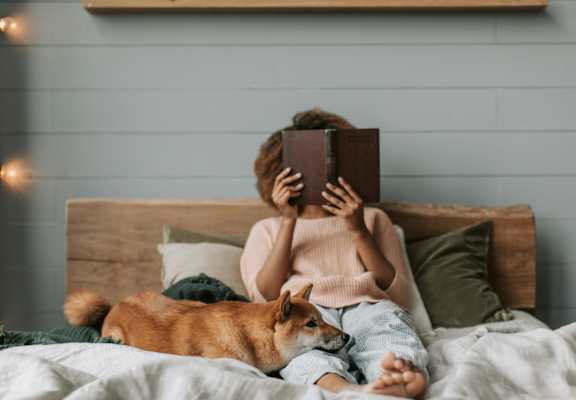 A person is reading a book while sitting on a bed with a dog.