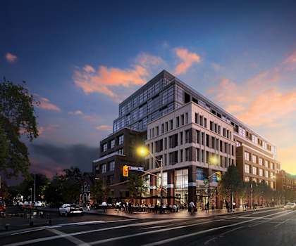 A modern building with glass windows is lit up at dusk with people walking on the sidewalk.