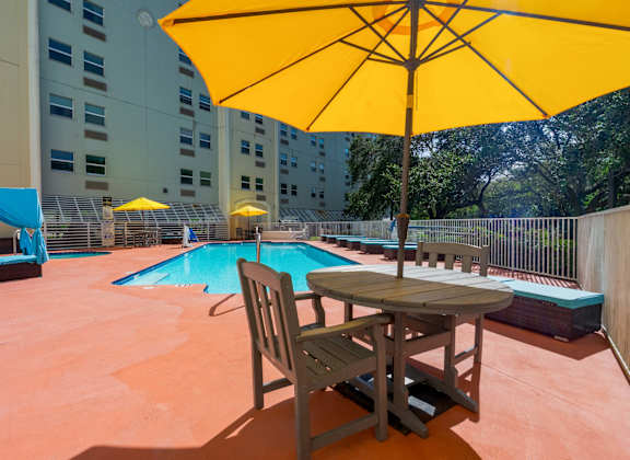 A yellow umbrella is on a table with a chair in front of a pool.