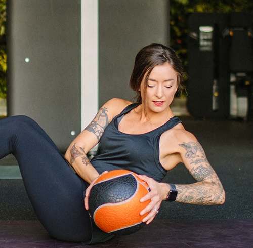 A woman with tattoos is sitting on a mat holding a medicine ball. at Bakery Living, Pennsylvania, 15206