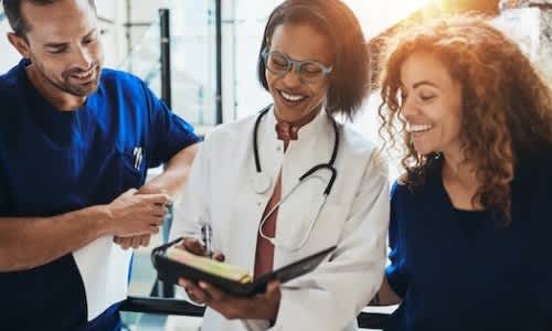 Three medical professionals, two wearing blue scrubs and one in a white coat, are engaged in a discussion over a tablet.
