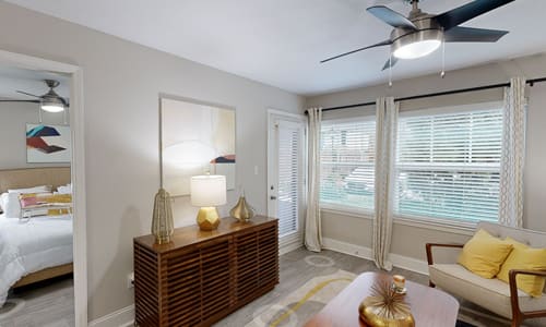A bedroom with a bed, a nightstand with a lamp, a table with a vase, a couch with a pillow, a fan, and a window with blinds.