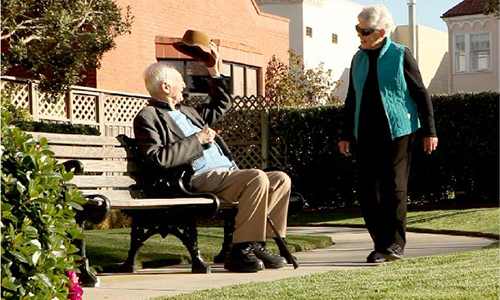 A man in a hat is sitting on a bench while another man walks by.