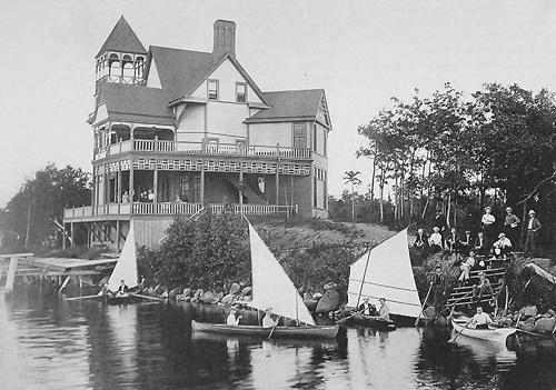 A black and white photo of a large house with a group of sailboats in front of it at The Barnum Apartments, White Bear Lake, Minnesota