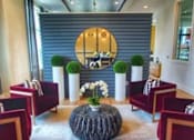 Thumbnail 11 of 17 - A room with a round mirror, a table with a vase of flowers, and chairs with red cushions.